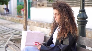 Canlı kıvırcık saçlı genç bir kadın şık ahşap bir bankta oturuyor, yanında şık bir çantayla akıllı telefonuna odaklanıyor. Dışarıdaki dingin ortam huzurlu anı güzelleştiriyor.