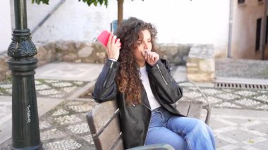 Güzel, kıvırcık saçlı, şık, genç bir kadın rahat rahat bir park bankında oturuyor. Etrafını saran gün ışığının keyfini çıkarırken akıllı telefonunu neşeyle tutar.