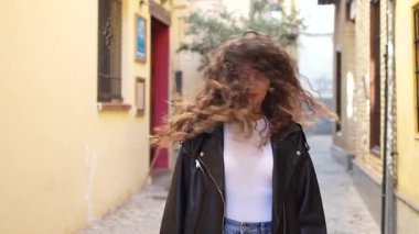 Güzel, kıvırcık saçlı, şık bir deri ceket giymiş genç bir kadın, canlı ve renkli bir şehir geçidinde güvenle duruyor, yüzü ışıl ışıl ışıl gülümserken.