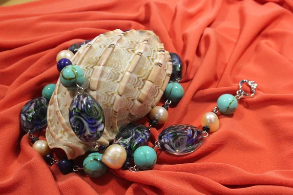 A large sea shell and a necklace of  necklace semi-precious stones - lapis lazuli, turquoise and pearls on a pink background.