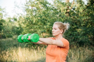 A woman doing dumbbell exercises outdoors. Outdoor sports. Green dumbbells in the athletes hands. 