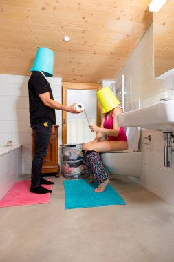 Kafasında kova olan bir adam tuvalette oturan karısına tuvalet kağıdını uzatıyor, kadın da kafasında kovayla oturuyor. Komik bir durum..