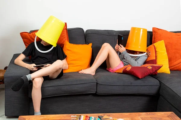 兄と妹は自宅のソファに座って バケツを頭にタブレットで遊んでいました コミュニケーションの欠如という概念 — ストック写真