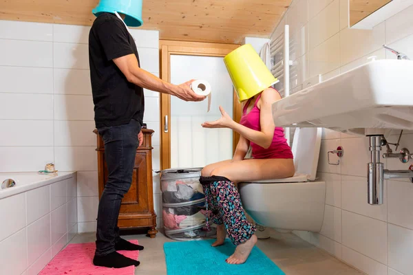 男人头上拿着桶 把厕纸递给坐在马桶上的妻子 妻子头上也拿着桶 有趣的情况 — 图库照片