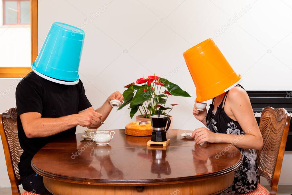 Young loving couple drinks coffee in the dining room with a bucket on their heads. Unreal situation and concept of non-communication