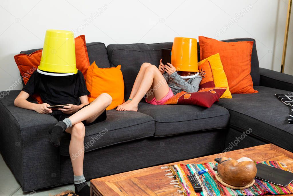 Brother and sister sitting on a sofa at home with a bucket on their heads playing with tablets. Concept of absence of communication