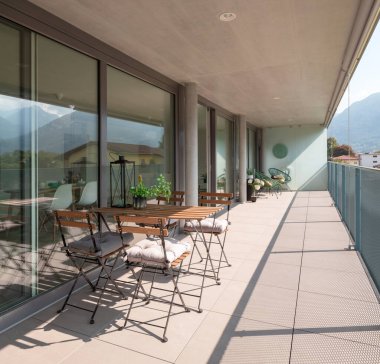 Ahşap masa ve sandalyeli modern ev terası. Masada bir fidan, bir şişe ve bir fener var. Yazın güneşli bir gün.