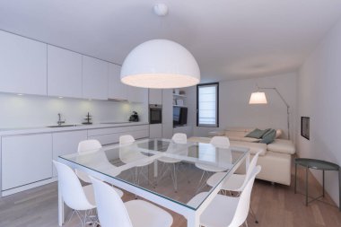 Geniş bir açık alan, cam masa, tasarımcı koltukları, beyaz mutfak ve rahat deri koltuklar. Beyaz duvarlar. İçeride kimse yok.