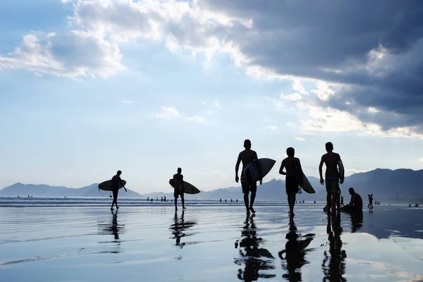Silhouette di surfisti irriconoscibili che trasportano la tavola da surf sulla spiaggia al tramonto, con una nuvola e specchi d'acqua e montagne sullo sfondo — Foto Stock