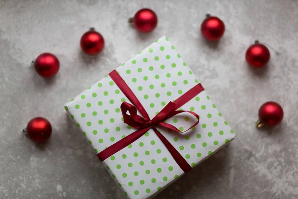 Χριστουγεννιάτικα στολίδια σε γκρι φόντο. Ένα λευκό και πράσινο κουτί με μπορντό φιόγκο που περιβάλλεται από μη εστιασμένη μπορντό Χριστουγεννιάτικες μπάλες, χριστουγεννιάτικα παιχνίδια και πέτρινο στολίδι. Άνω όψη. — Φωτογραφία Αρχείου