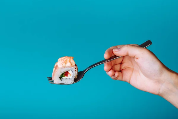 Hand holding fresh kunsan sushi roll with a fork, isolated on blue background. How to eat sushi without chopsticks.