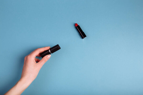 Lady's hand holding closed red lipstick isolated on blue background near to opened lipstick.