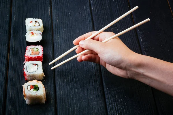 La mano femenina toma palillos rollos de sushi con palillos chinos. Acostado sobre una tabla de madera negra. Vista desde arriba —  Fotos de Stock