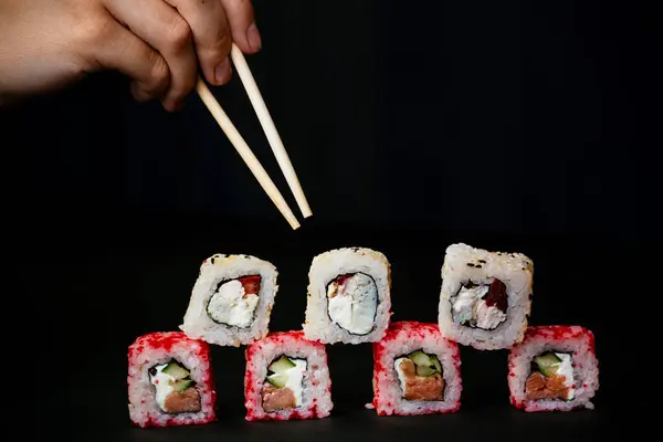 Mão feminina leva pauzinhos rolos de sushi com pauzinhos chineses. Deitado numa tábua de madeira preta. Vista de cima — Fotografia de Stock