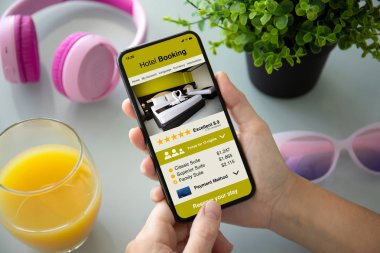 female hands holding phone with app hotel booking on the screen above the table with headphones