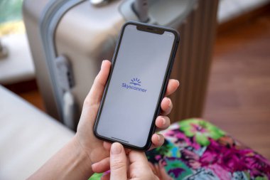 Alanya, Türkiye - 12 Haziran 2020: Uygulamalı uçak bileti satış hizmeti havayolları Skyscanner ile iPhone 11 ile el ele tutuşan kadın ekranda ve bagajda.