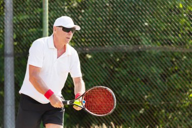 Fiziksel olarak iyi durumda yaşlı bir tenisçi güneşli bir günde açık havada tenis oynar.