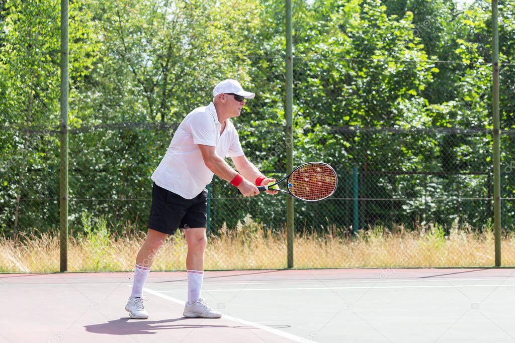An elderly man in good physical shape plays tennis on an outdoor court on a sunny day