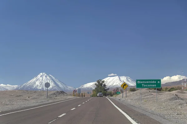 Lunga strada dritta con cartello giallo che conduce verso una montagna innevata nel deserto di Atacama, Cile — Foto Stock