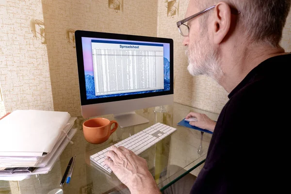 髭のコンピューター上のスプレッドシートに取り組んでいる中年の男性 フォルダーのファイル オレンジ カップがテーブルに置かれる — ストック写真