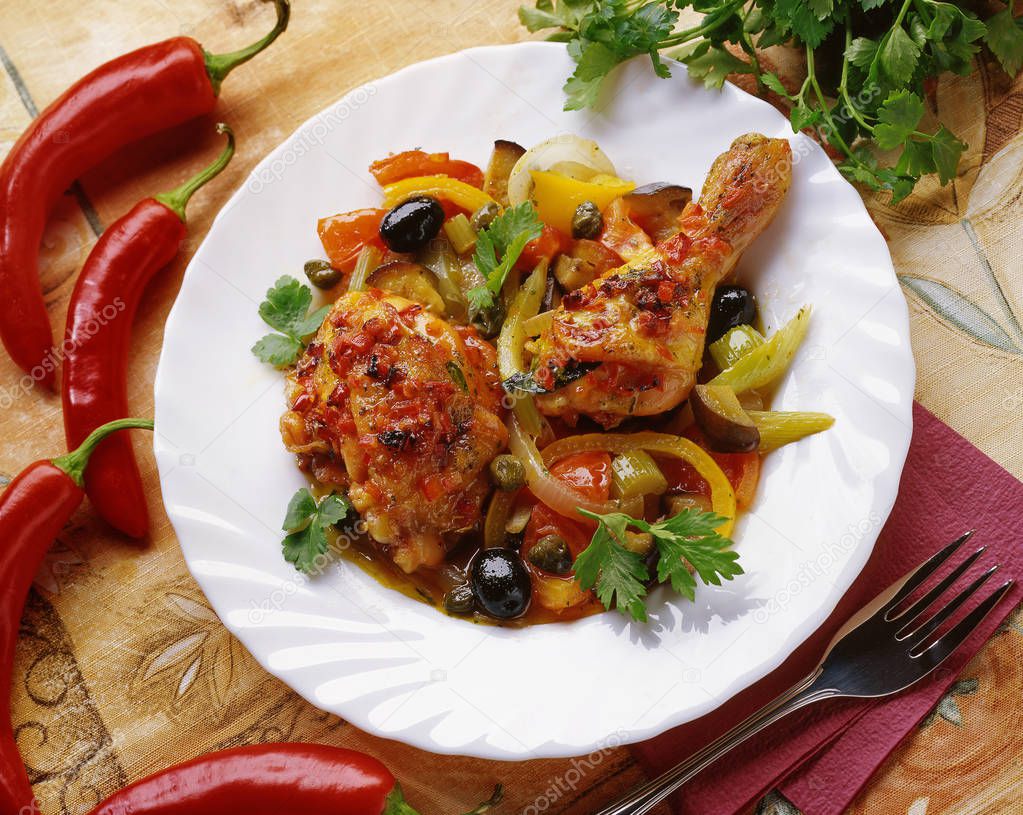 Stewed chicken with vegetables: onion, pepper, olives, celery, eggplant, peas, red pepper on a white plate, served on a table