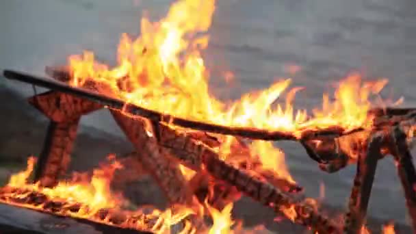 Geen geluid. Extreme close-up twintig tweede video van een brandende picknicktafel aan het einde van een feestje door een waterlichaam. Zomer vakantie leuk als de tafel valt. Razende vlammen, intense hitte en rook. — Stockvideo