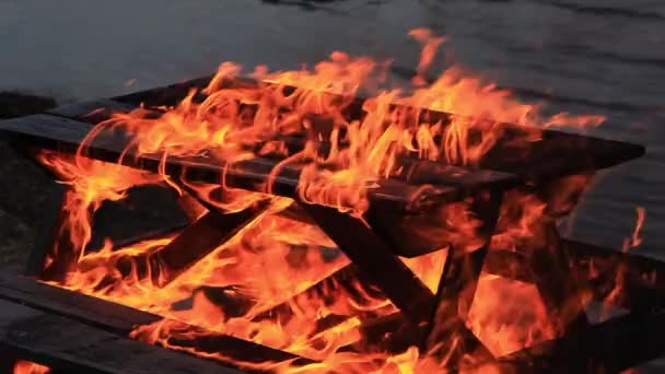 Suono. Intensi tre 3 secondi di bellissime fiamme arancioni e rosse che inghiottono un tavolo da picnic in legno vicino a un specchio d'acqua la sera presto nella notte d'estate . — Video Stock