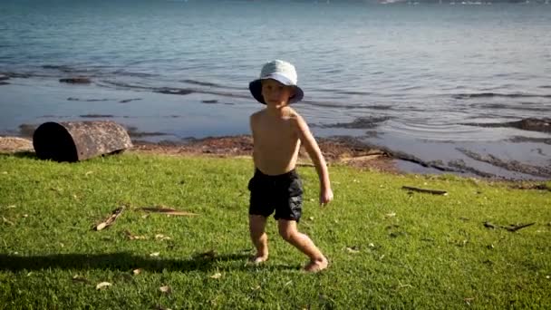 Young boy in shorts and bucket hat on the grassy shore of a lake at the height of summer showing his version of how to do "The Floss" — Stock Video