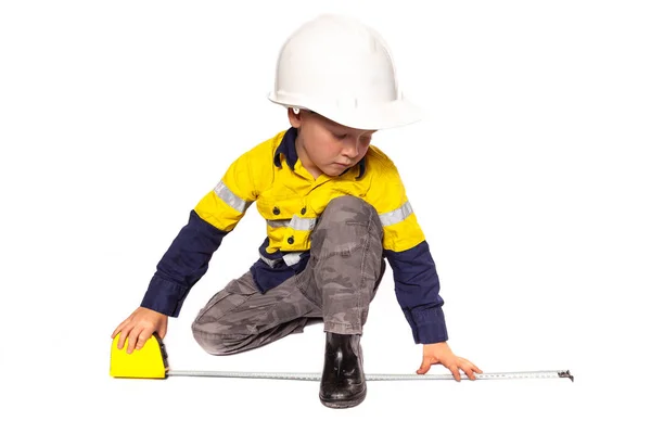 Junger blonder kaukasischer Junge, der als Bauarbeiter in gelb-blauem Hi-viz-Hemd, Stiefeln, weißem Hut und Maßband eine Rolle spielt. Stockfoto