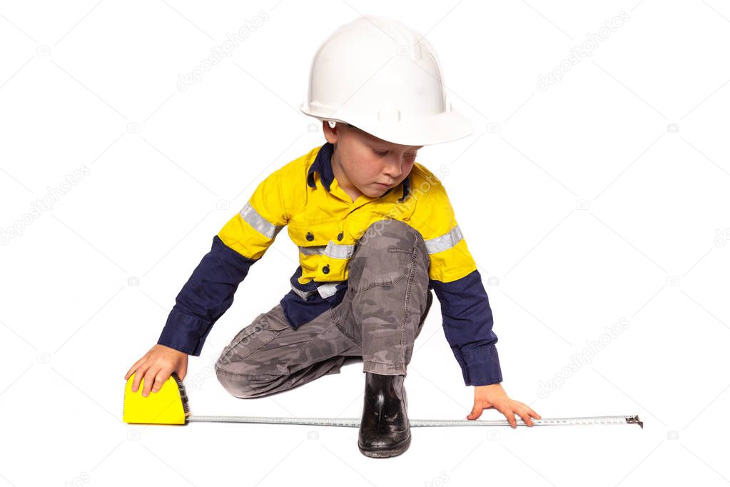Young blond caucasian boy measuring something role playing as a construction worker in a yellow and blue hi-viz shirt, boots, white hard hat, and tape measure.