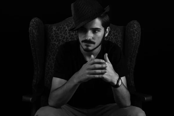 Close up of young adult male looking sinister or contemplative. Monotone, black and white for dramatic effect, dark and moody series. Concept image for magician in wingback chair unhappy and scheming.