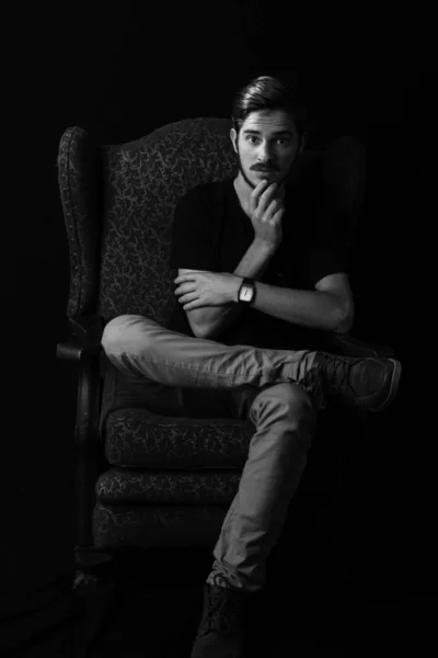 Young adult male sitting in vintage wingback chair with hand on chin, surprise expression. Monotone, black and white for dramatic effect. Concept for facial expressions for acting school.