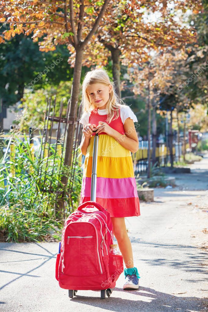 Cute 8 years old girl going to school, carry red backpack with wheels