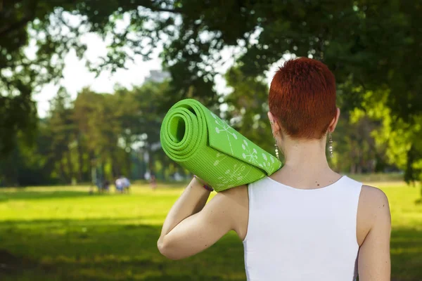 Exercise in nature, recreation in beautiful sunlit park. Beautiful female fitness instructor holding an exercise mat above her arm, active sport lifestyle