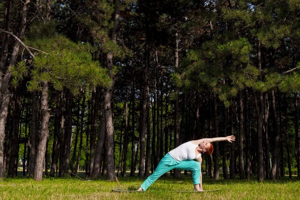 瑜伽教练户外 Utthita Trikonasana 红发女郎在公园做瑜伽 后面的树木 阳光普照 — 图库照片