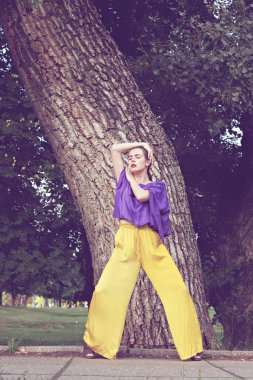 Genç ve güzel kadın moda model parkta ağaç tarafından duran kamera için poz