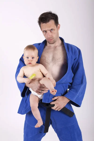 Athletic karate man in blue kimono with black belt standing, holding a baby boy and looking at camera
