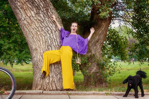 Genç ve güzel kadın parkta ağaç tarafından kamera ayakta için poz süre dalga geçmeyi