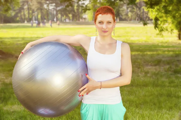 Pilates recreation in nature - Portrait of Cheerful young redhead woman in fitness wear exercising with  fitness ball in park