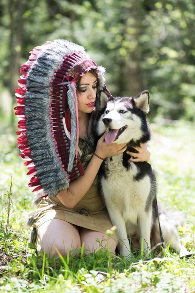Young woman in traditional red indian warbonnets posing with black husky in green meadow