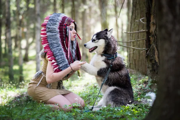 Young woman in traditional red indian warbonnets posing with black husky in green forest