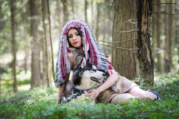 Young woman in traditional red indian warbonnets posing with black husky in green forest