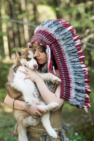 Young woman in traditional red indian warbonnets posing with brown husky puppy in green forest