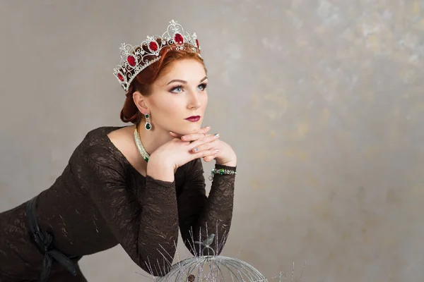 elegant woman in crown with rubies leaning on bird cage on grey stone background