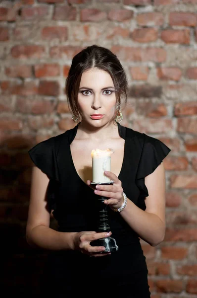 Young elegant woman in black dress holding candle and posing on brick wall background