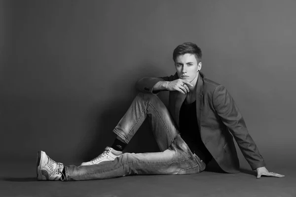 Young serious man in black t-shirt and stylish blazer sitting and posing on grey background