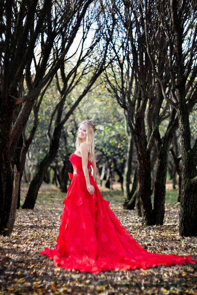 Young woman in elegant red dress and jewel crown posing in autumn forest