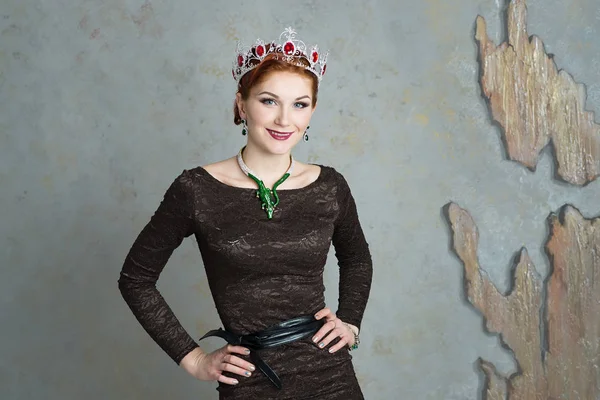 Portrait of elegant woman in crown with rubies and necklace with green gemstones on grey stone background