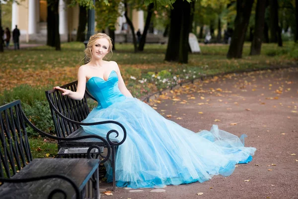 Blonde woman in sky blue fairy dress sitting on metal bench in autumn park