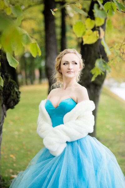 Elegant blonde woman in fairy dress and white short fur coat posing park